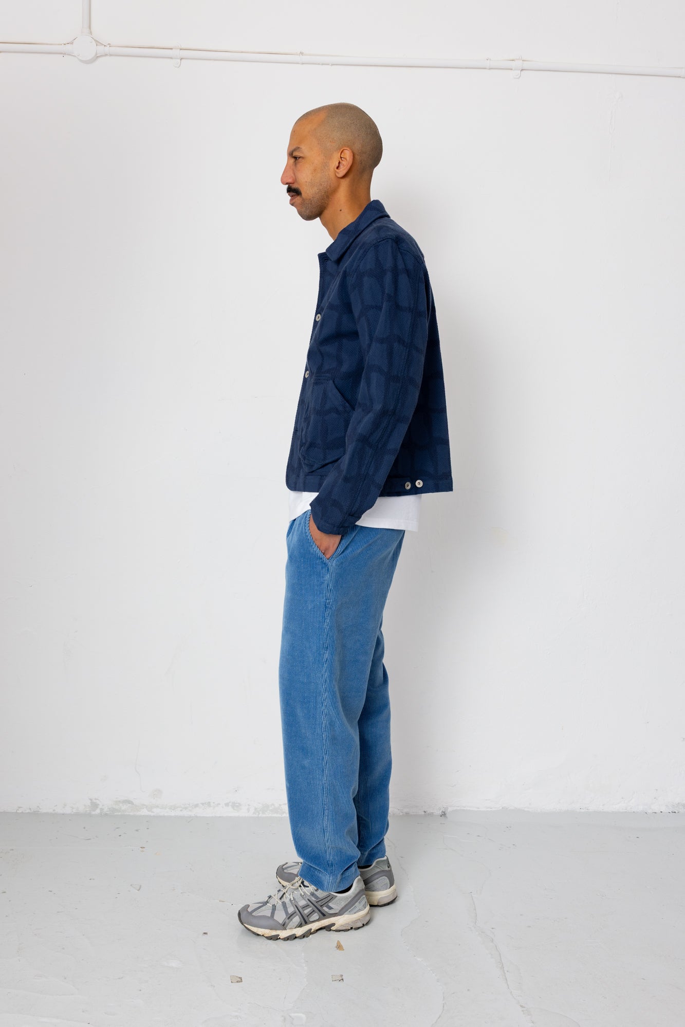 Man wearing a blue jacket and jeans standing against a white wall.