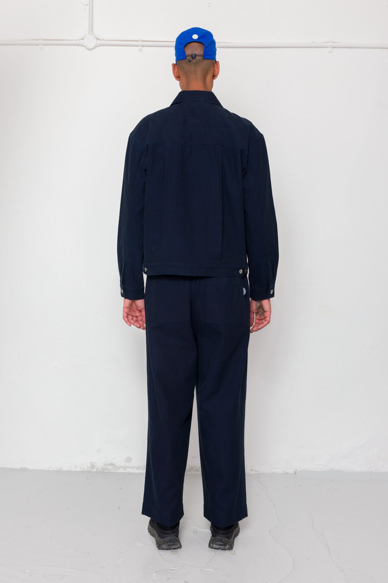 Person wearing a navy jacket & trousers with a blue cap against a white background