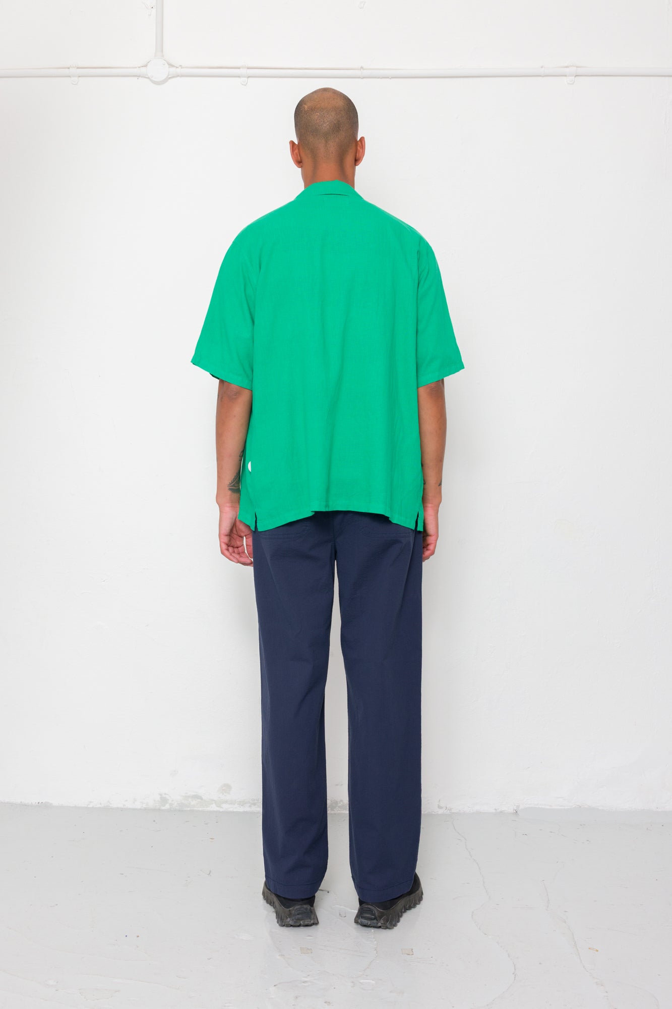 Person wearing a green shirt and navy pants standing against a white wall.
