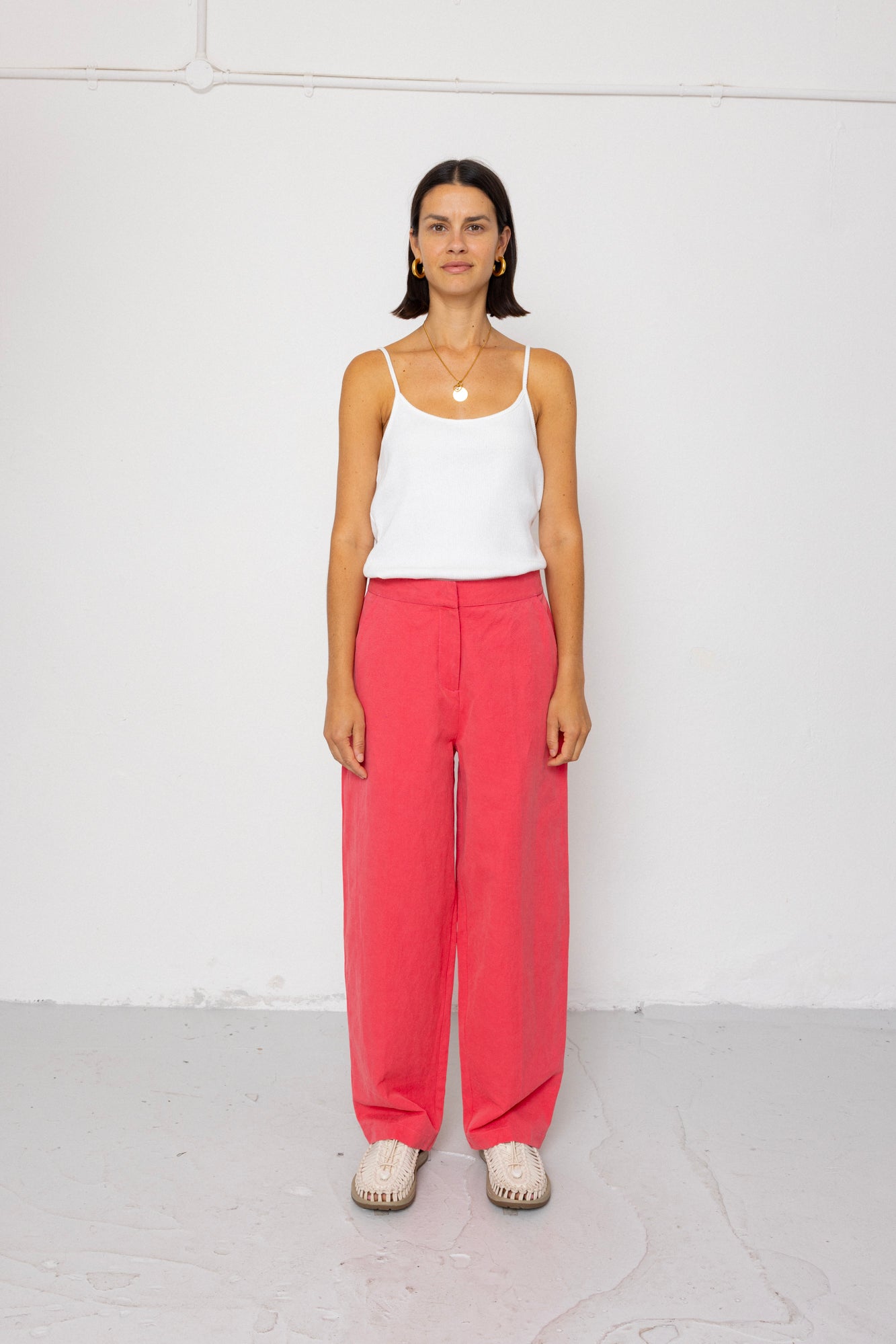 Woman wearing a white tank top and coral pants against a white background