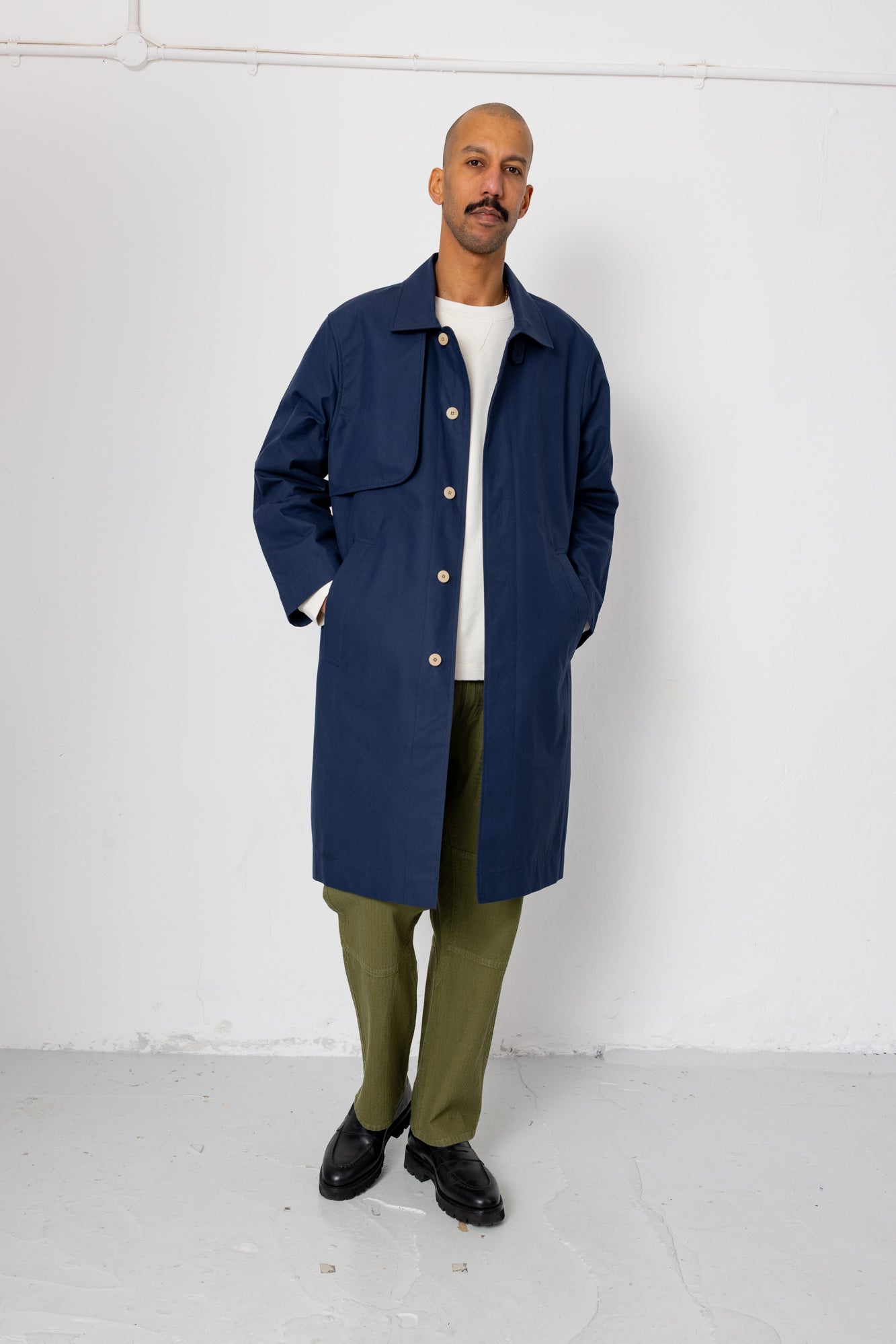 Man wearing a navy coat, white shirt, green pants, and black shoes against a white background