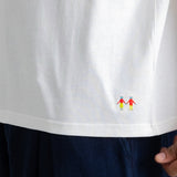 Close-up of a white short-sleeve T-shirt worn with dark navy pants, showing a small colorful embroidered graphic near the lower right hem. A hand is visible at the edge holding the shirt.
