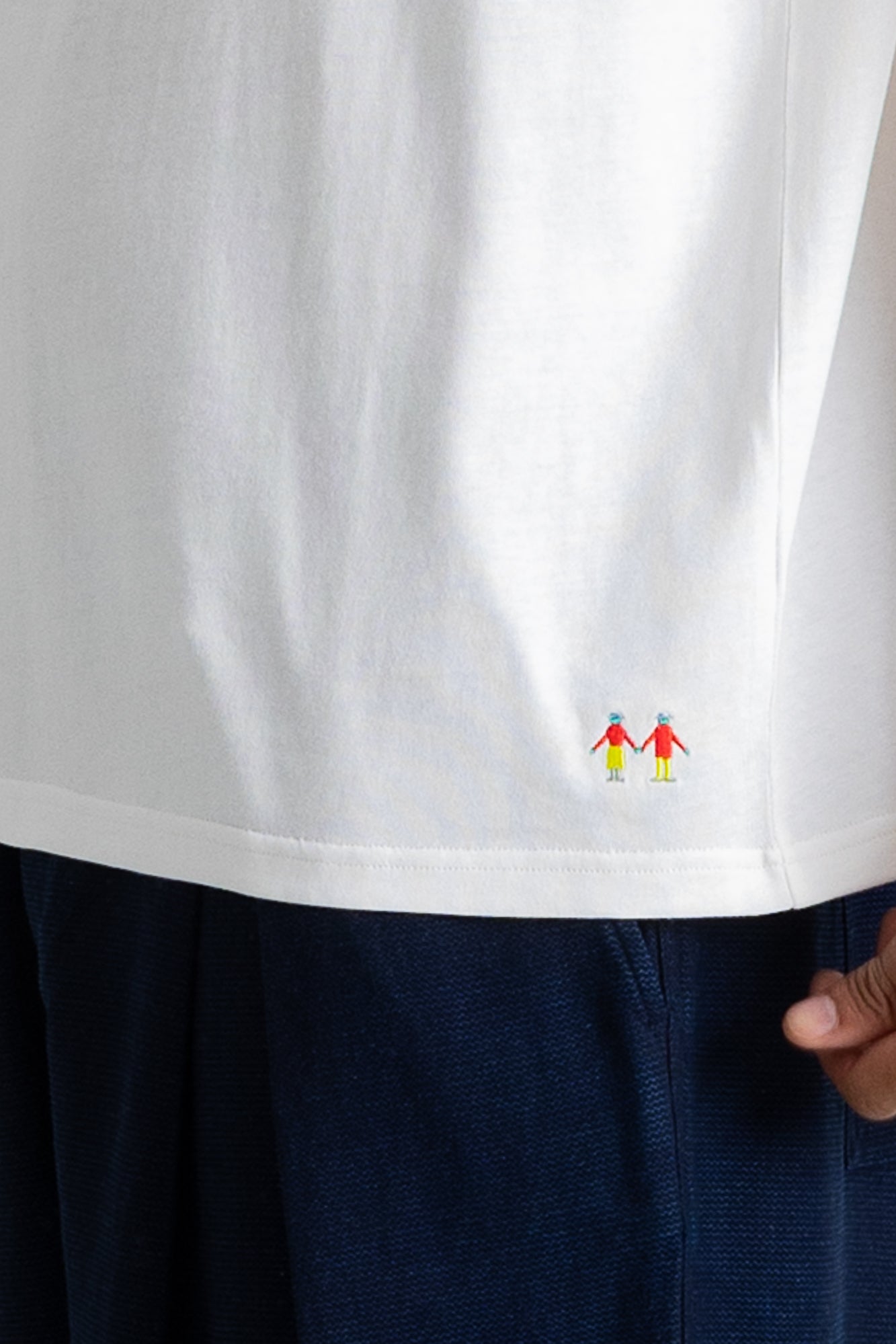 Close-up of a white short-sleeve T-shirt worn with dark navy pants, showing a small colorful embroidered graphic near the lower right hem. A hand is visible at the edge holding the shirt.