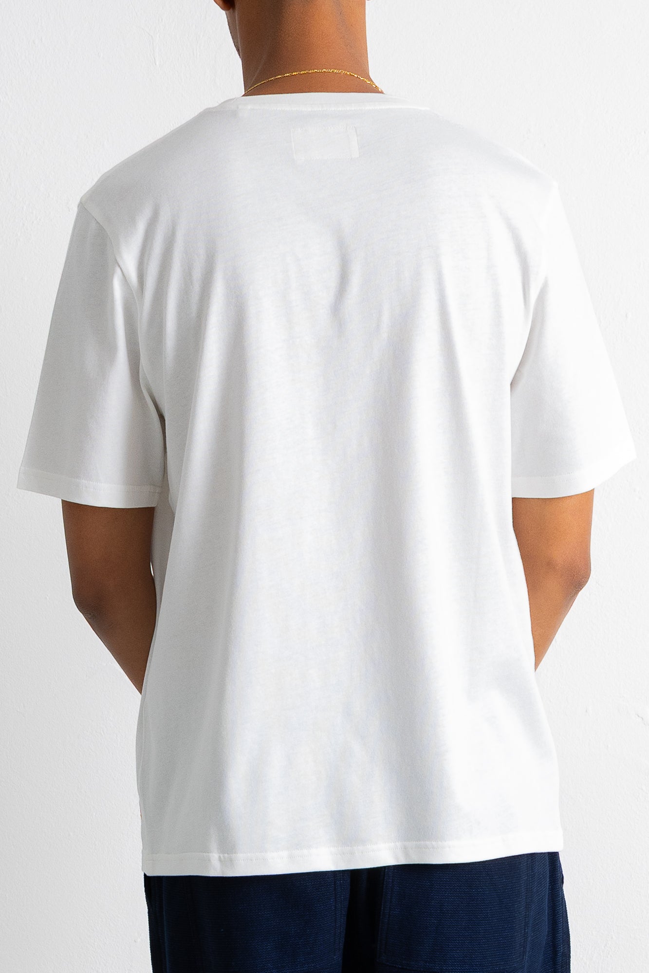 A person shown from the back wearing a plain white short-sleeve crew-neck T-shirt and dark navy pants against a light gray studio background.