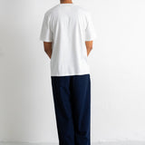 A man viewed from behind wearing a white short-sleeve t-shirt and dark navy wide-leg trousers, standing against a plain white wall.