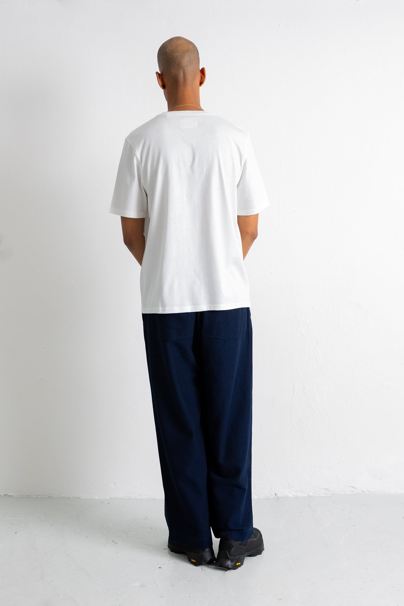 A man viewed from behind wearing a white short-sleeve t-shirt and dark navy wide-leg trousers, standing against a plain white wall.