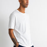 A man wearing a plain white short-sleeve crewneck t-shirt and dark navy pants, standing in profile against a light gray studio background.