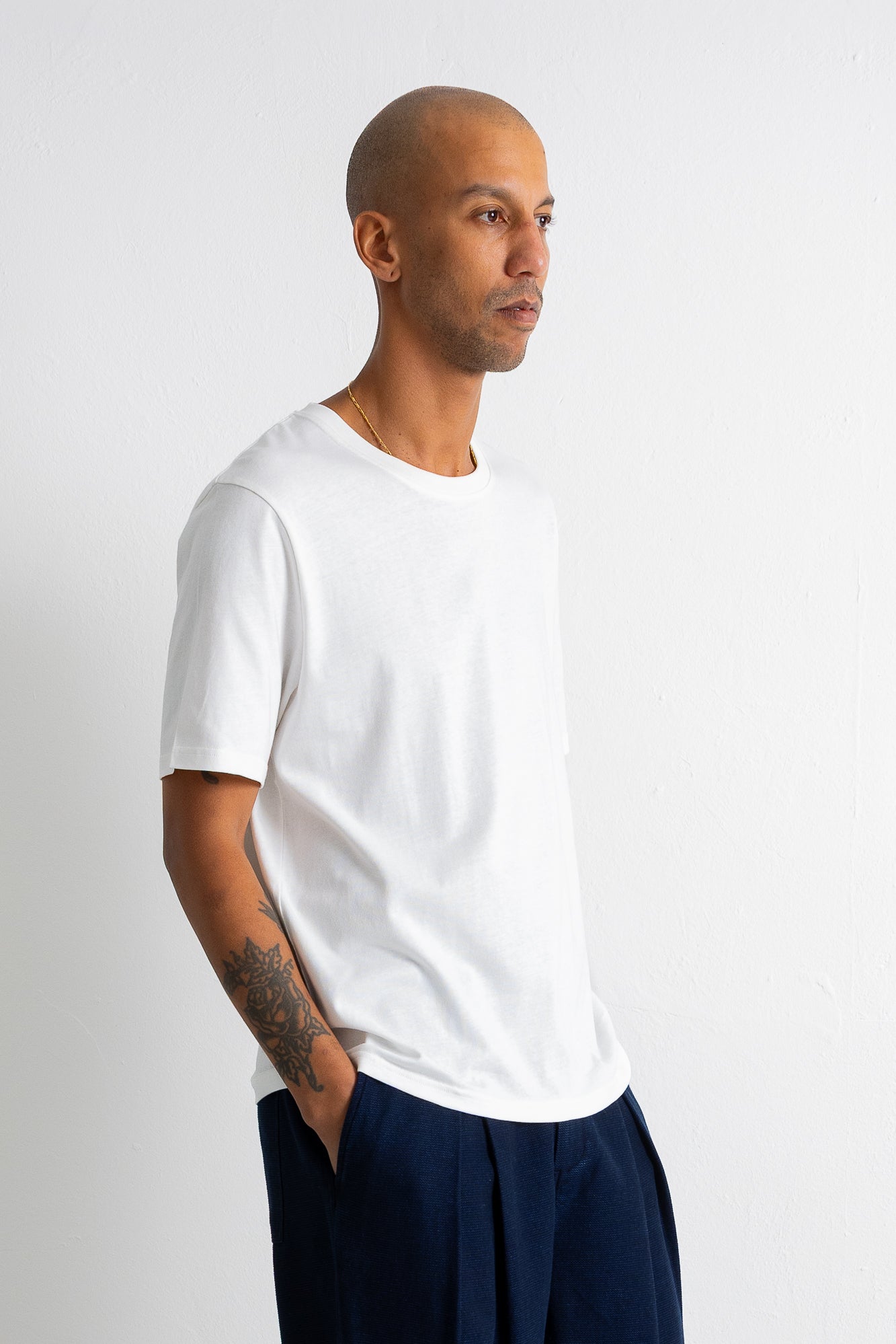 A man wearing a plain white short-sleeve crewneck t-shirt and dark navy pants, standing in profile against a light gray studio background.