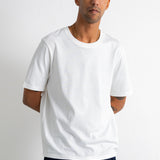 A man wearing a plain white short-sleeve crewneck t-shirt and dark navy pants, standing against a light gray studio background.
