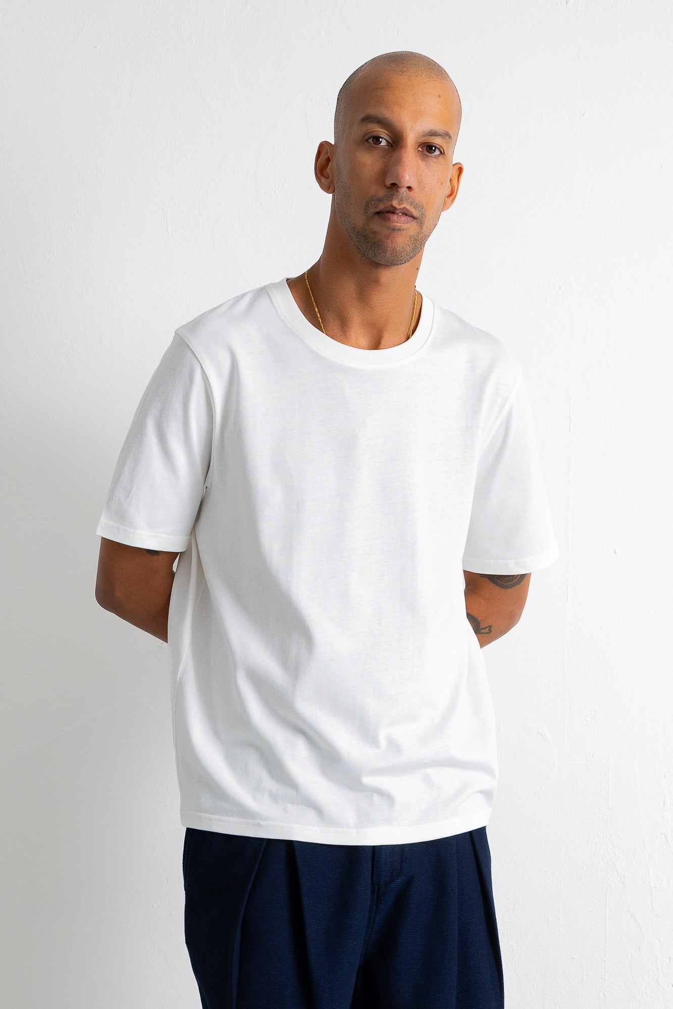 A man wearing a plain white short-sleeve crewneck t-shirt and dark navy pants, standing against a light gray studio background.