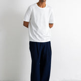 A man wearing a plain white short-sleeve crewneck t-shirt and loose dark navy blue pants, standing against a clean white studio background.