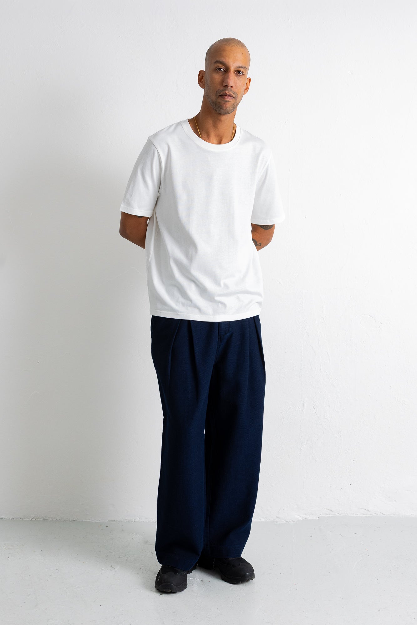 A man wearing a plain white short-sleeve crewneck t-shirt and loose dark navy blue pants, standing against a clean white studio background.