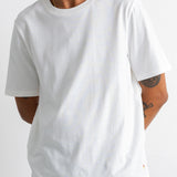 A man wearing a plain white short-sleeve crewneck t-shirt and dark navy pants, cropped from the mouth down against a light gray studio background.