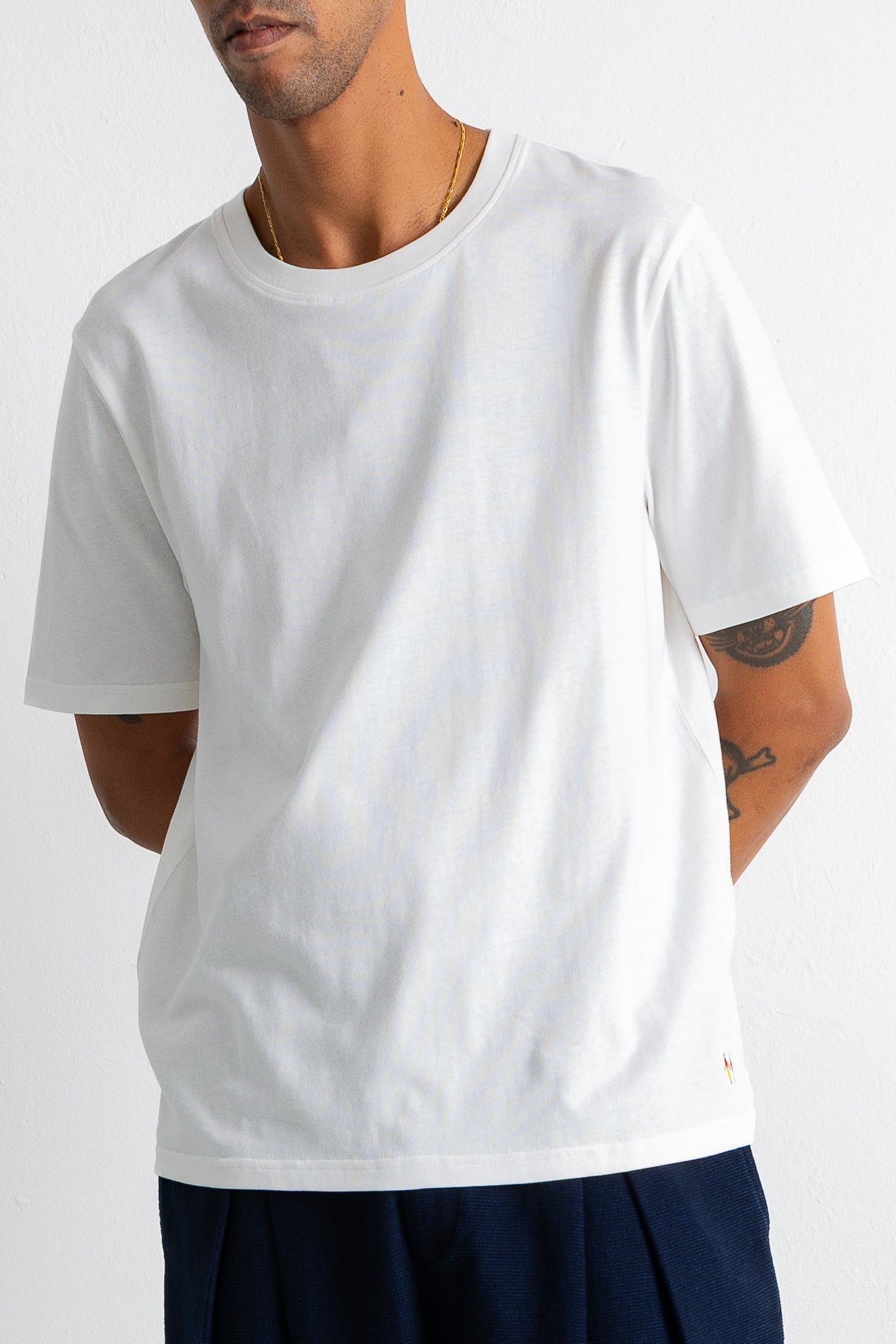 A man wearing a plain white short-sleeve crewneck t-shirt and dark navy pants, cropped from the mouth down against a light gray studio background.