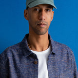 A man wearing a muted green baseball cap with a small embroidered logo, a textured blue button-up shirt over a white t-shirt, set against a solid blue background.