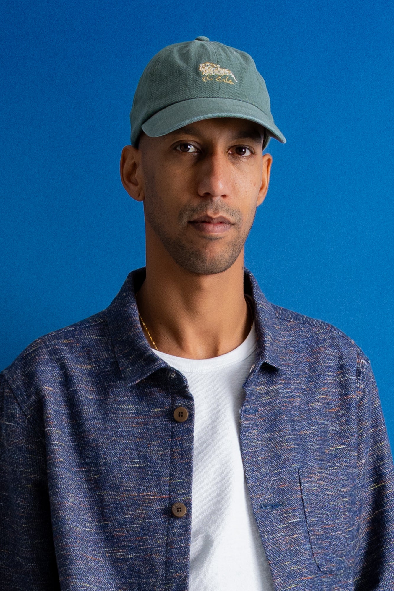 A man wearing a muted green baseball cap with a small embroidered logo, a textured blue button-up shirt over a white t-shirt, set against a solid blue background.