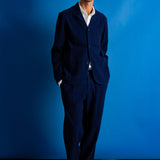 A man wearing a dark navy blue two-piece suit with a white collared shirt underneath, standing against a solid blue studio backdrop.