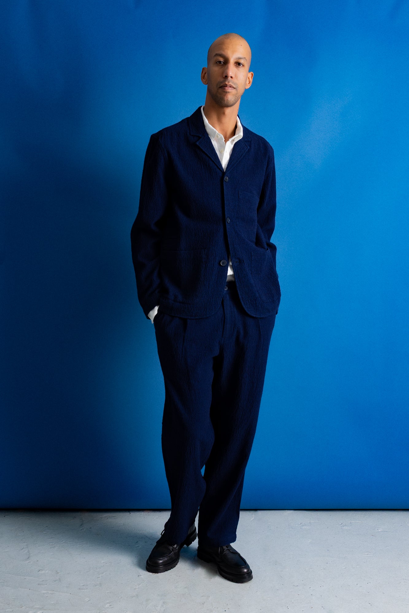 A man wearing a dark navy blue two-piece suit with a white collared shirt underneath, standing against a solid blue studio backdrop.