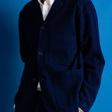 A man wearing a dark navy blue two-piece suit with a white collared shirt underneath, standing against a solid blue studio backdrop.