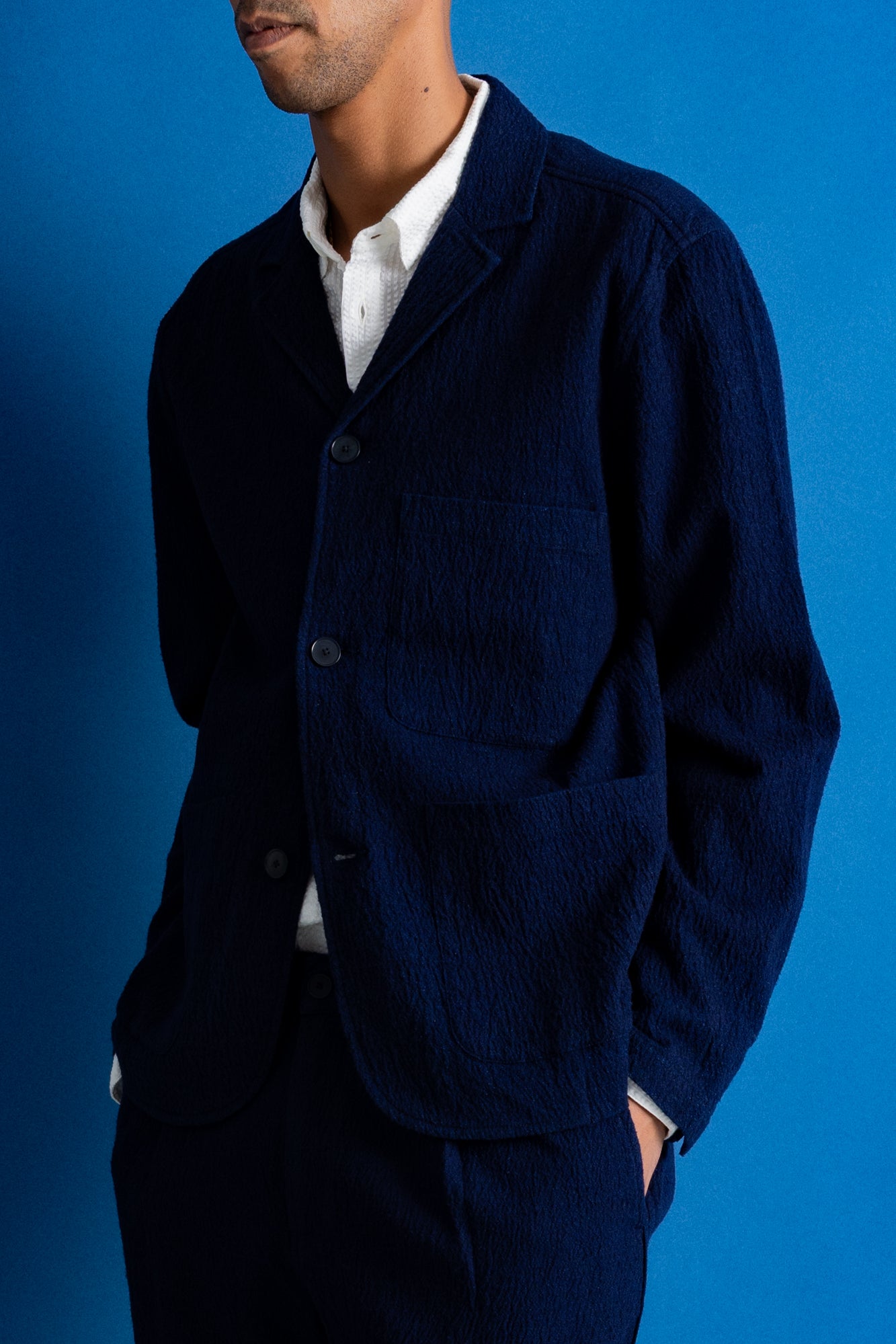 A man wearing a dark navy blue two-piece suit with a white collared shirt underneath, standing against a solid blue studio backdrop.