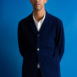 A man wearing a dark navy blue two-piece suit with a white collared shirt underneath, standing against a solid blue studio backdrop.