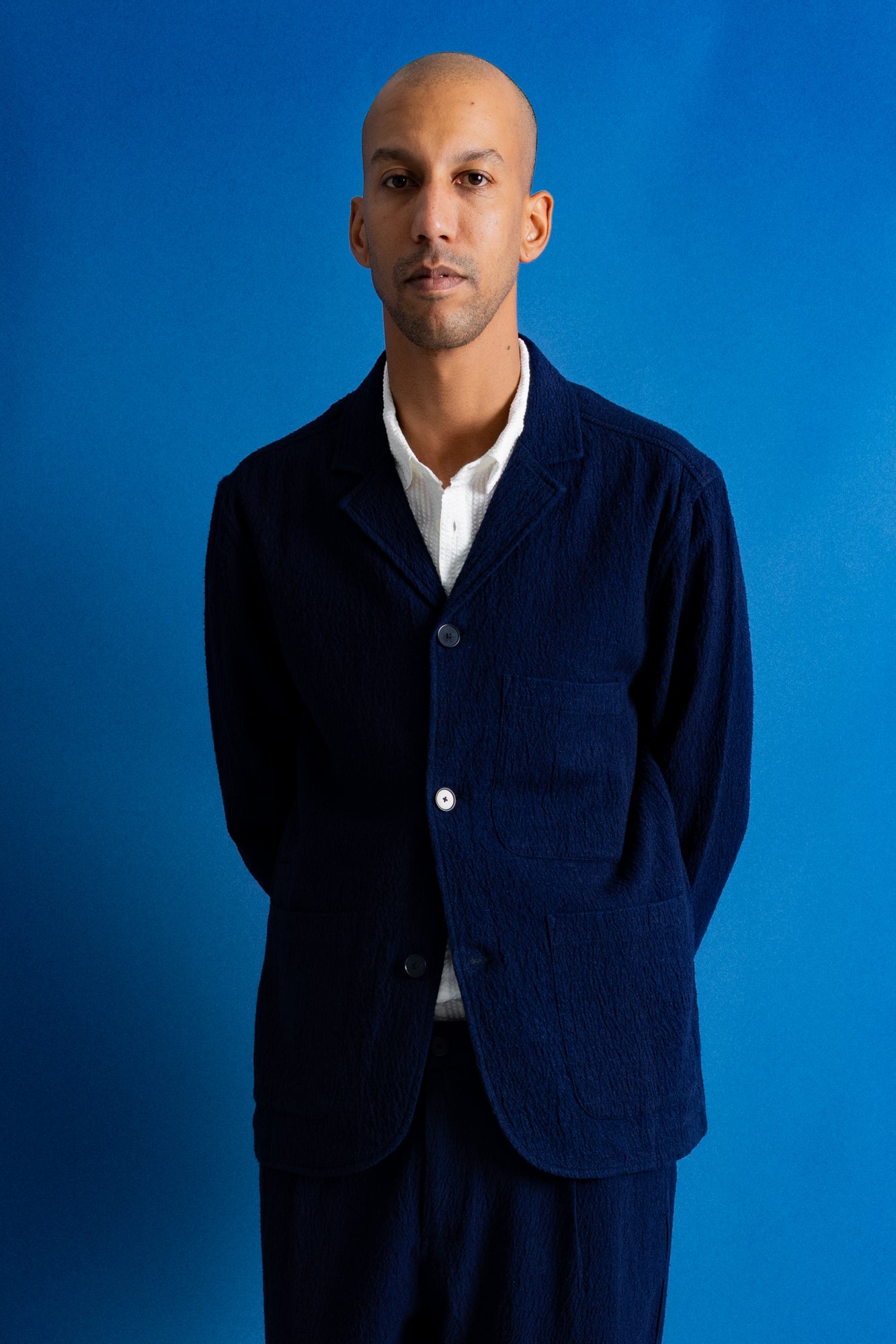 A man wearing a dark navy blue two-piece suit with a white collared shirt underneath, standing against a solid blue studio backdrop.