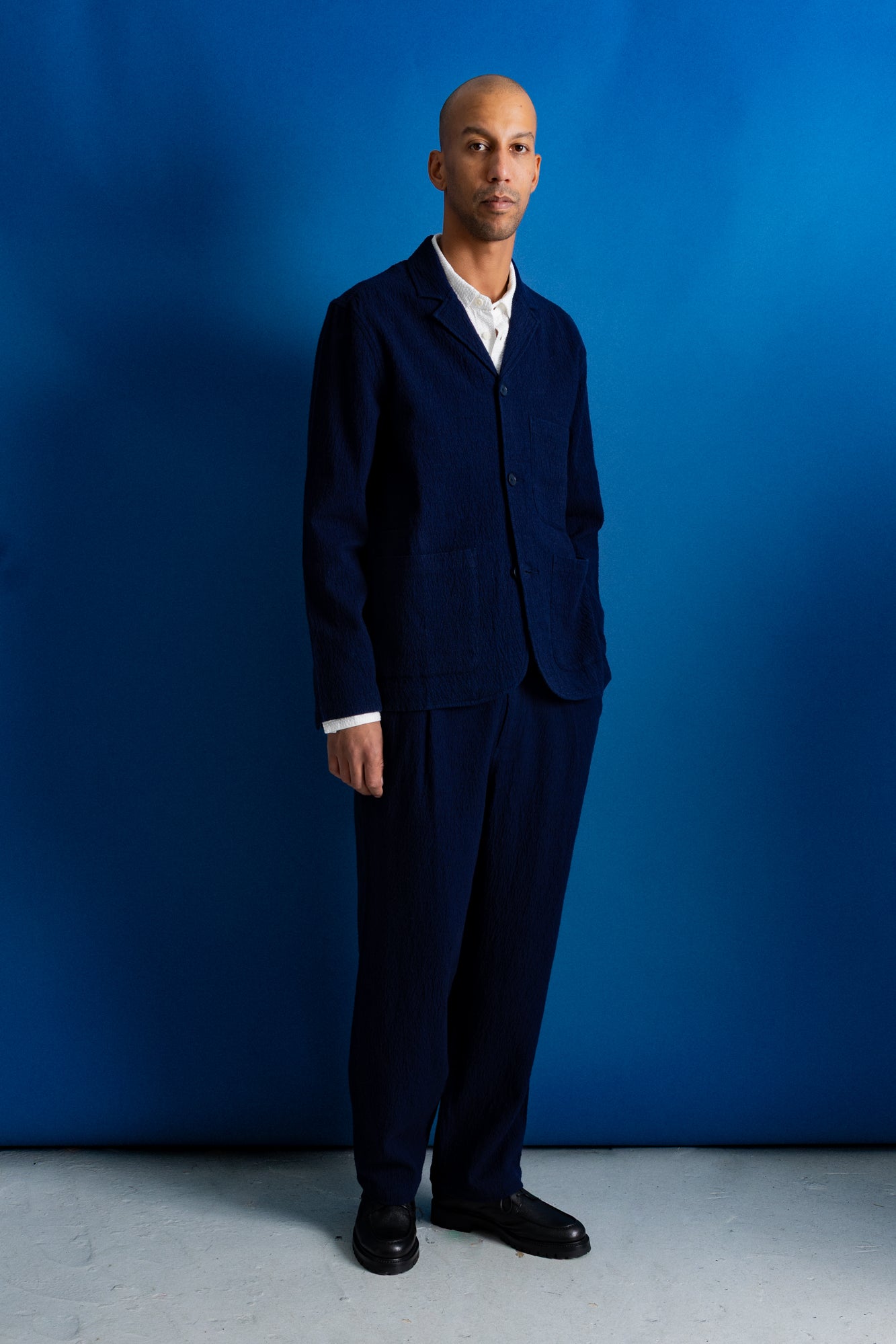A man wearing a dark navy blue two-piece suit with a white collared shirt underneath, standing against a solid blue studio backdrop.