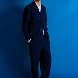 A man wearing a dark navy blue two-piece suit with a white collared shirt underneath, standing against a solid blue studio backdrop.