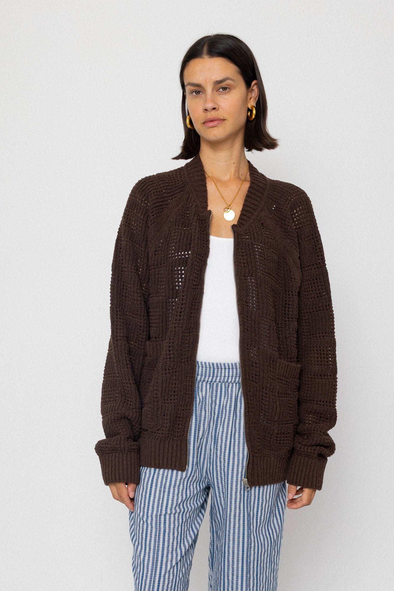 Person wearing a brown cardigan over a white top and blue striped pants on a plain background