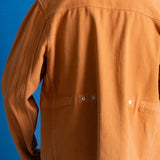A person shown from the back wearing a rust-orange utility jacket over a white t-shirt, set against a solid blue background.