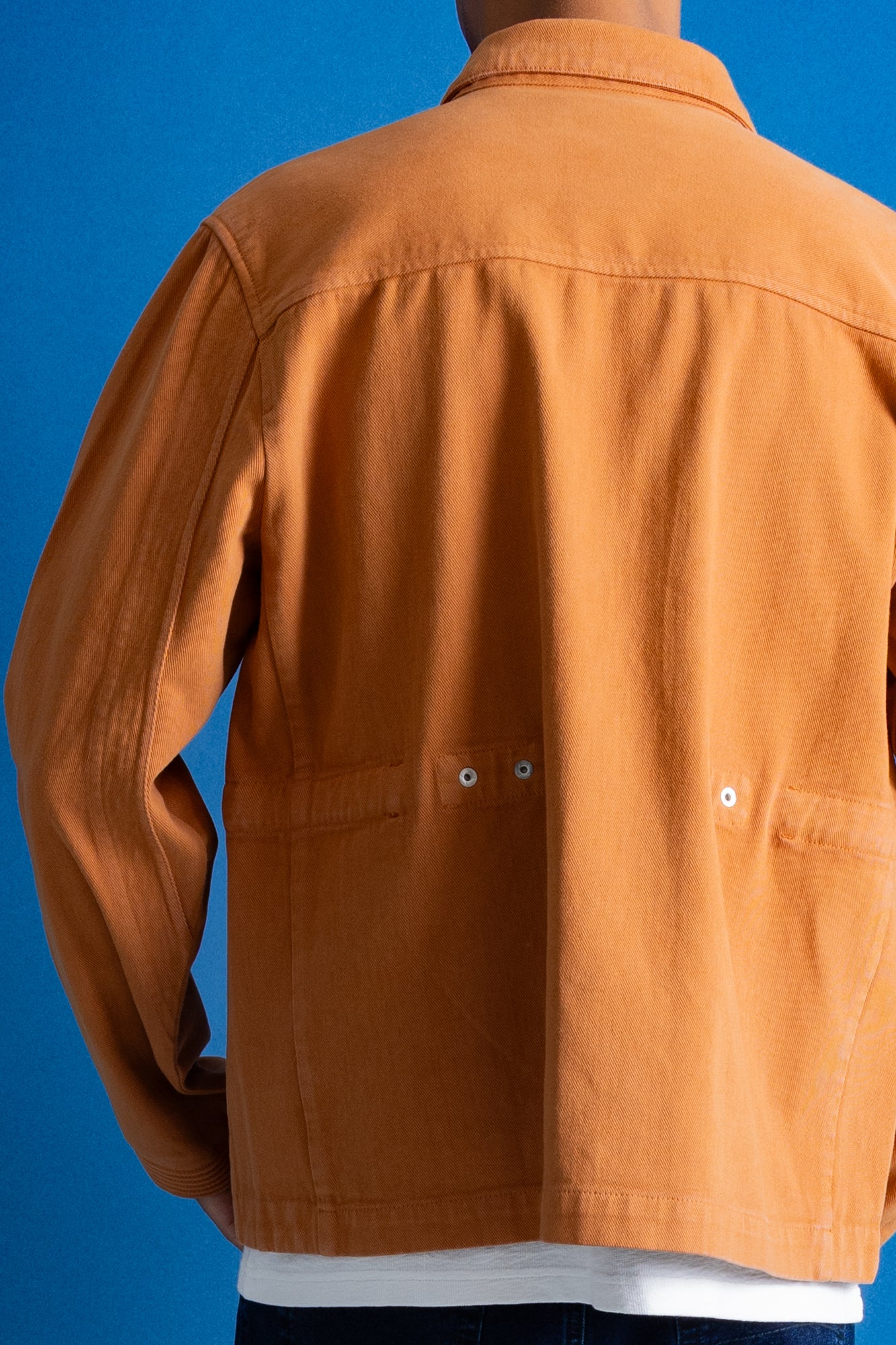 A person shown from the back wearing a rust-orange utility jacket over a white t-shirt, set against a solid blue background.