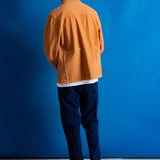 A man viewed from behind wearing an orange overshirt, dark navy trousers, and black shoes, standing against a solid blue studio backdrop.