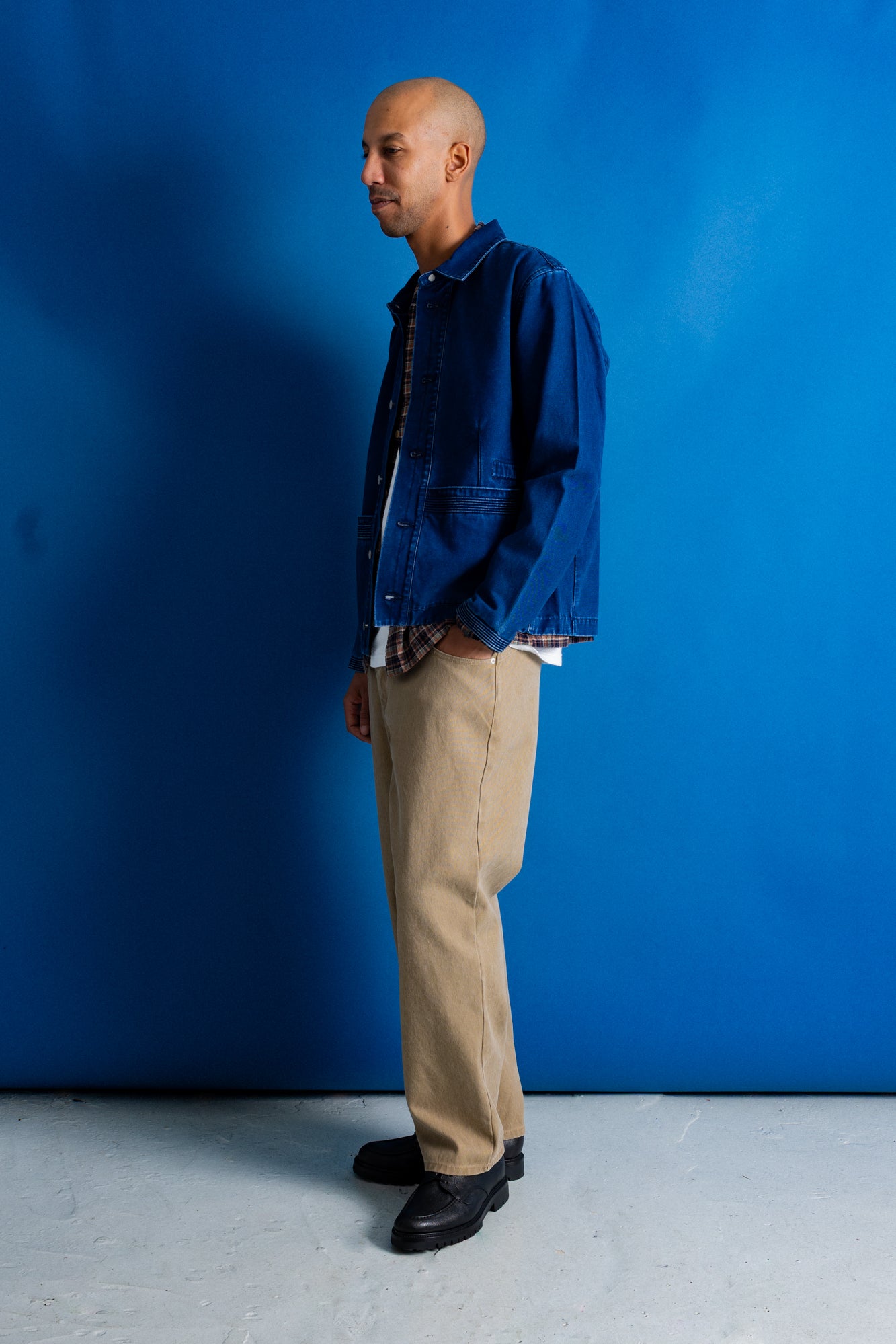 A man stands in profile wearing a blue denim jacket over a dark shirt, beige trousers, and black shoes against a bright blue studio backdrop.