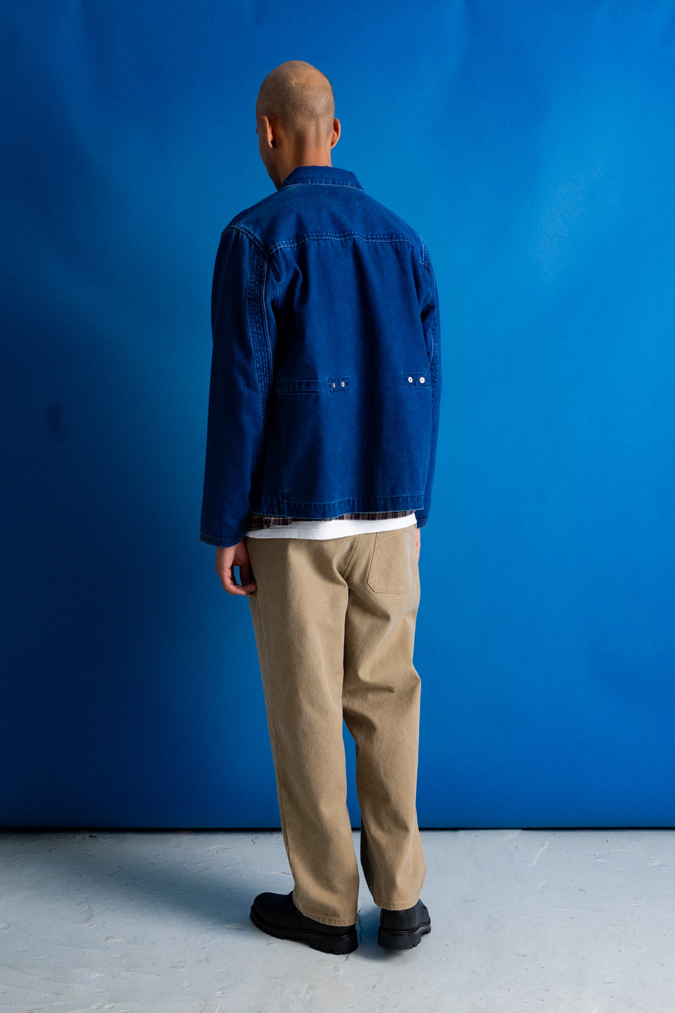 A man viewed from behind wearing a dark blue denim jacket, beige trousers, and black shoes, standing against a solid blue studio backdrop.