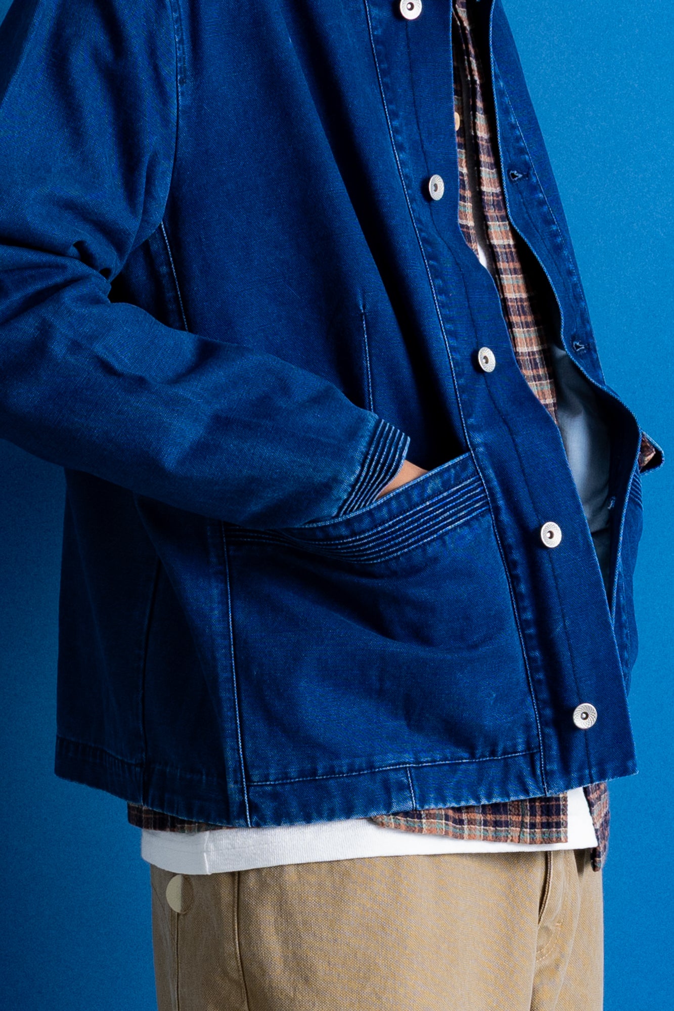 A close up of a pocket detail on a navy jacket, beige trousers, and checkered shirt, standing against a solid blue studio backdrop.