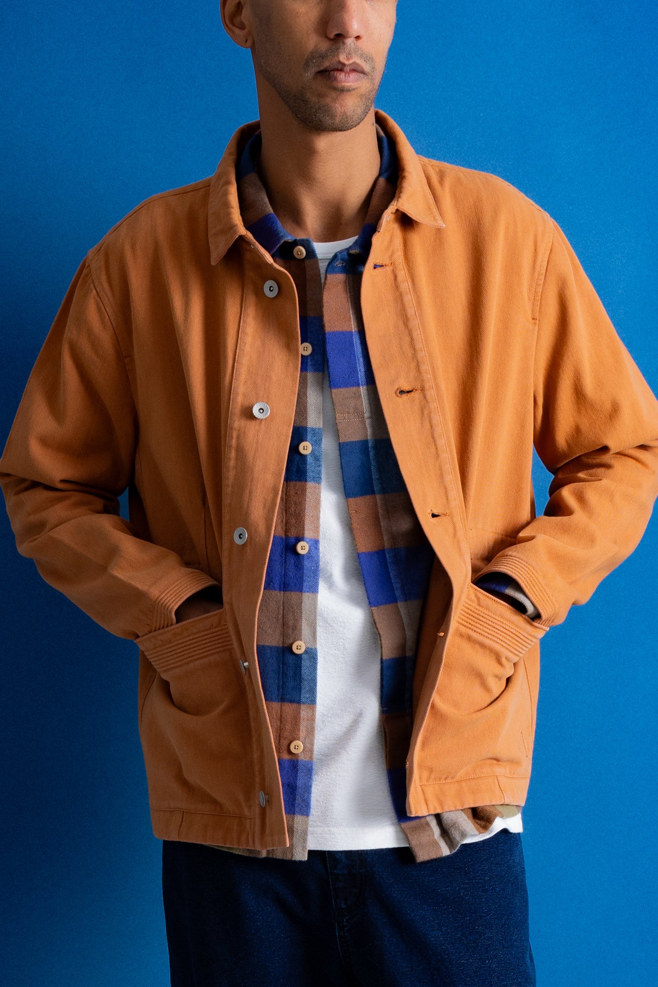 A man wearing a camel-colored overshirt jacket over a blue and brown plaid button-down shirt and a white t-shirt, set against a solid blue background.