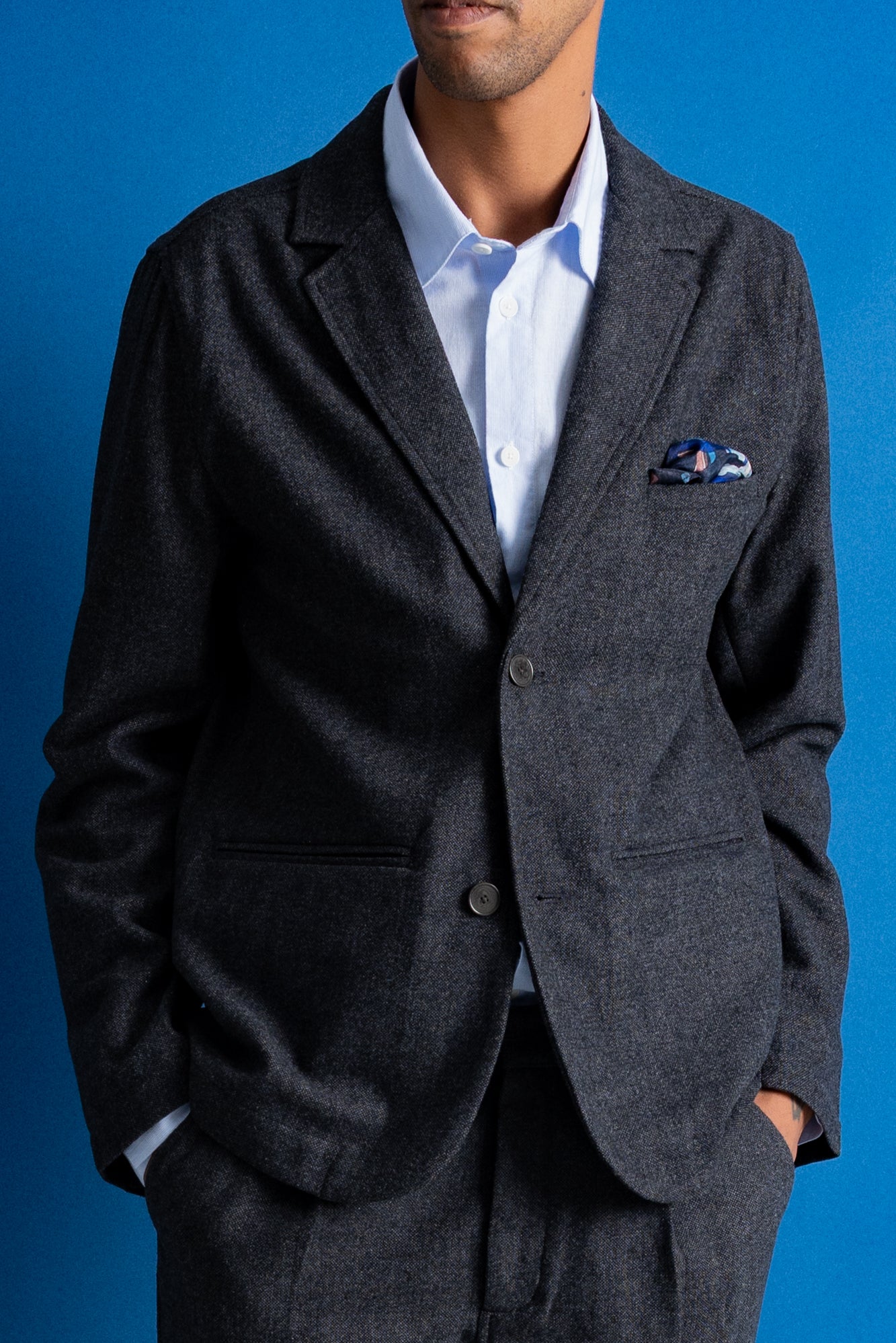 A man wearing a dark gray textured blazer over a light blue dress shirt, with a patterned pocket square in the breast pocket, set against a solid blue background.