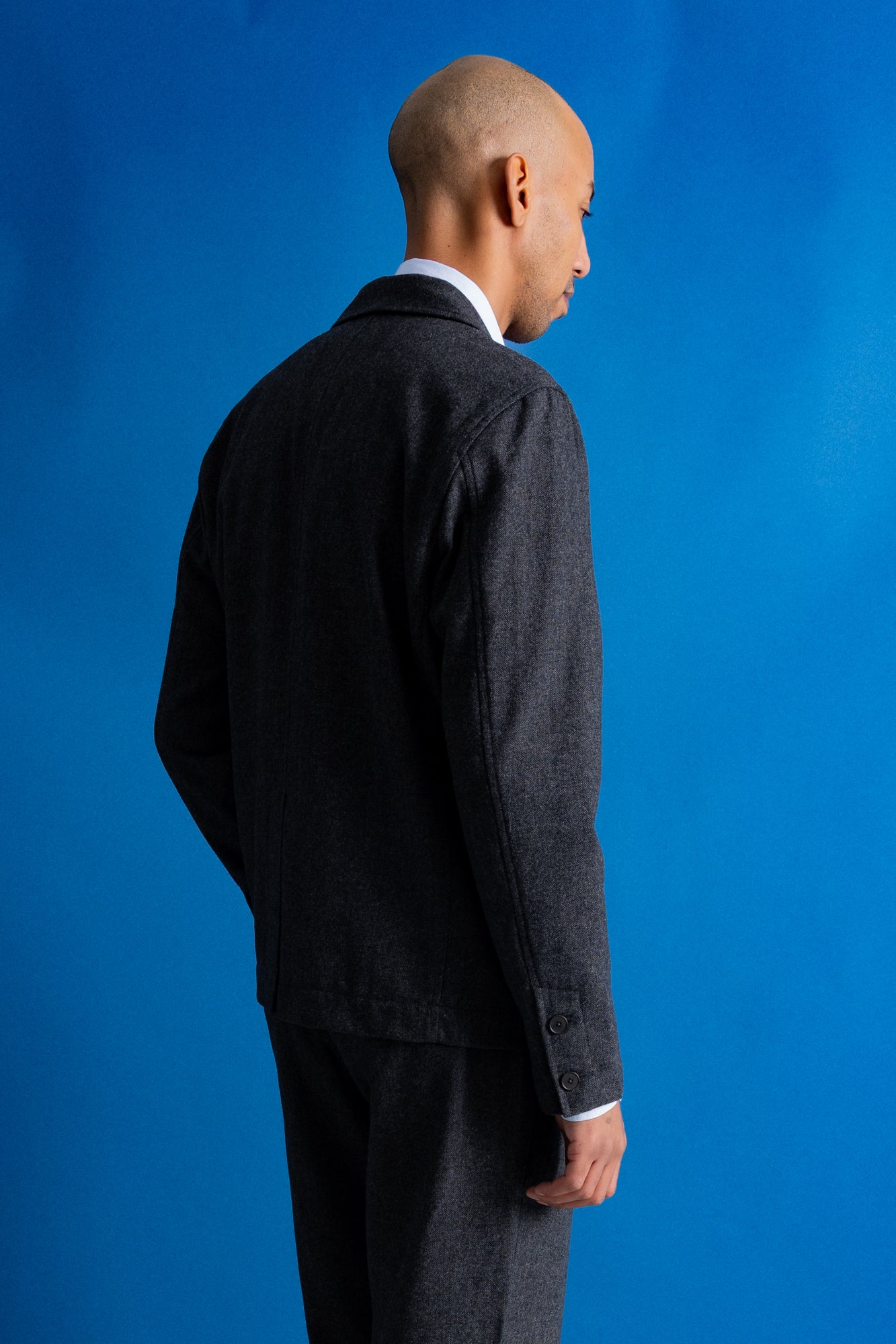 A man wearing a dark charcoal gray textured blazer and matching trousers over a white collared shirt, standing against a solid blue studio background.