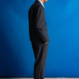A man in a dark gray two-piece suit, white dress shirt, and black dress shoes stands in profile against a bright blue studio backdrop.