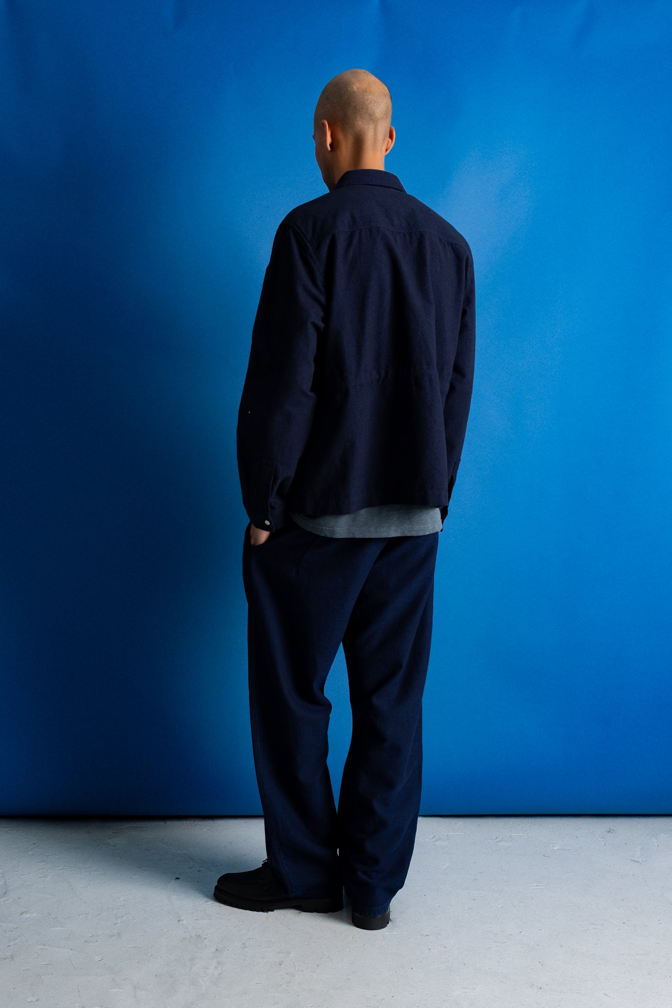 Man facing away from the camera wearing navy trousers and a grey green t-shirt underneath a moleskin navy jacket, standing against a bright blue background.