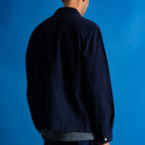 Man facing away from the camera wearing navy trousers and a grey green t-shirt underneath a moleskin navy jacket, standing against a bright blue background.