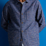 A man wearing a textured blue button-up overshirt jacket over a white t-shirt, set against a solid blue background.