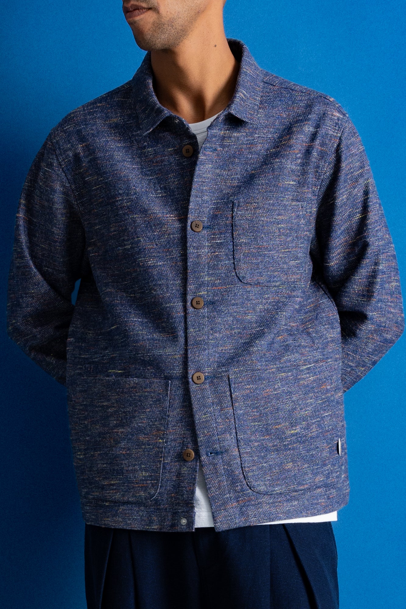 A man wearing a textured blue button-up overshirt jacket over a white t-shirt, set against a solid blue background.