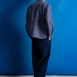 A man stands with his back to the camera wearing a textured blue and gray button-up shirt, dark navy trousers, black shoes, and a green cap, set against a solid blue studio backdrop.