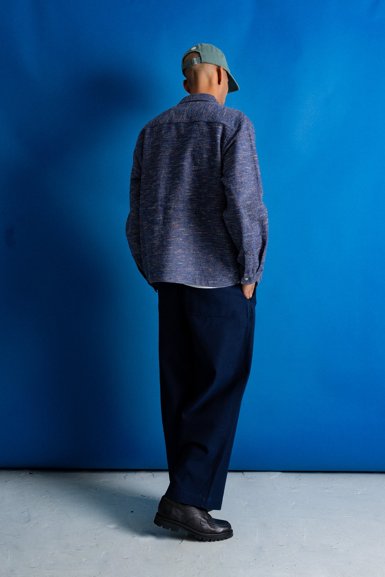 A man stands with his back to the camera wearing a textured blue and gray button-up shirt, dark navy trousers, black shoes, and a green cap, set against a solid blue studio backdrop.