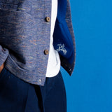 Close-up of a person wearing a textured blue tweed jacket over a white shirt and navy trousers, with a blue pocket showing a white embroidered logo, set against a solid blue background.