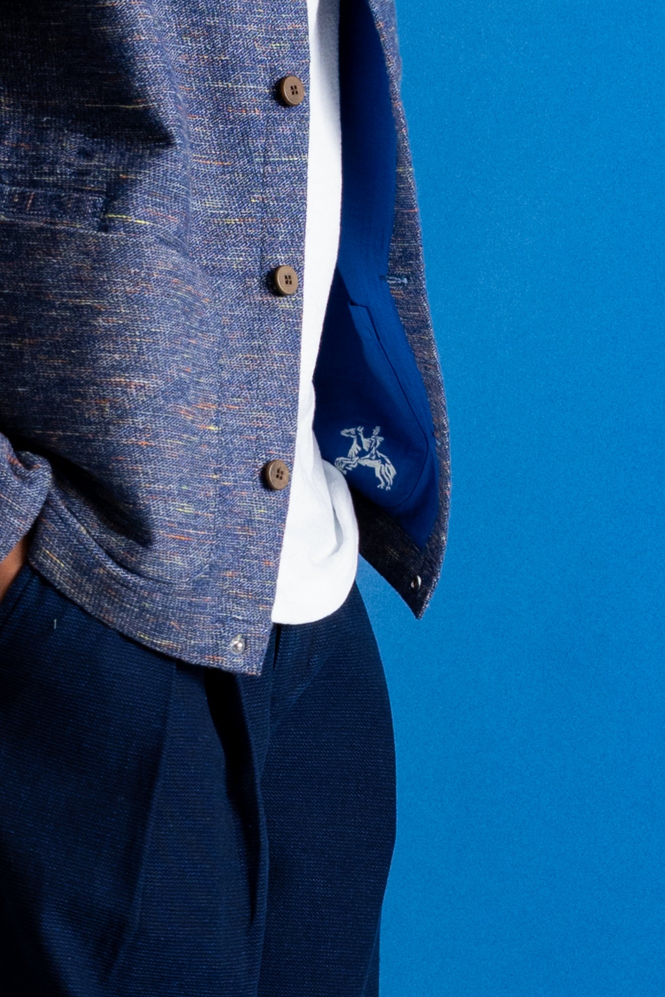 Close-up of a person wearing a textured blue tweed jacket over a white shirt and navy trousers, with a blue pocket showing a white embroidered logo, set against a solid blue background.