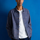 A man wearing a muted green baseball cap with a small embroidered logo, a textured blue button-up shirt over a white t-shirt, set against a solid blue background.