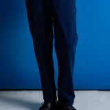 A person from the waist down wearing dark navy blue relaxed-fit trousers, a white t-shirt, and black leather shoes, standing against a vibrant blue wall.