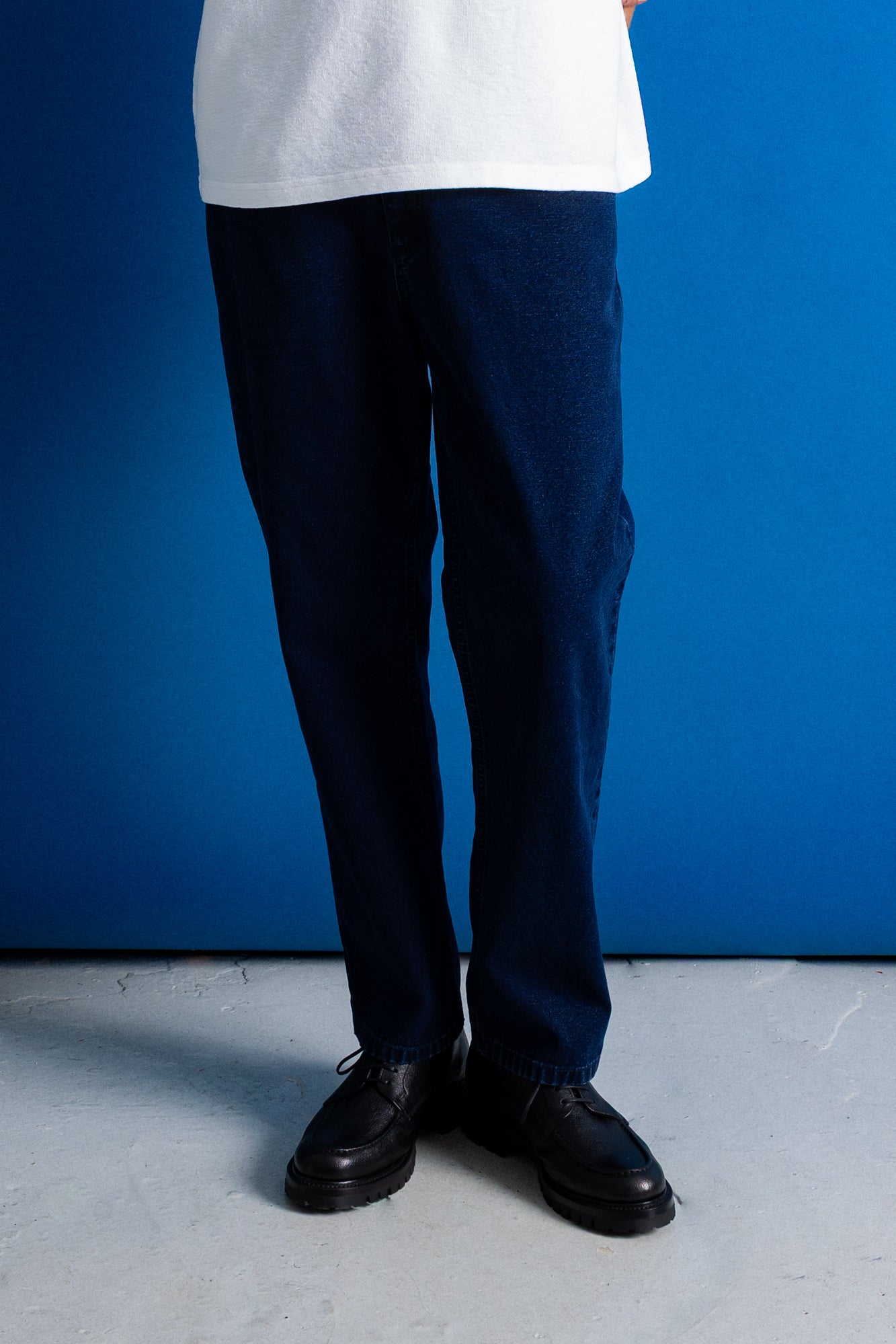 A person from the waist down wearing dark navy blue relaxed-fit trousers, a white t-shirt, and black leather shoes, standing against a vibrant blue wall.